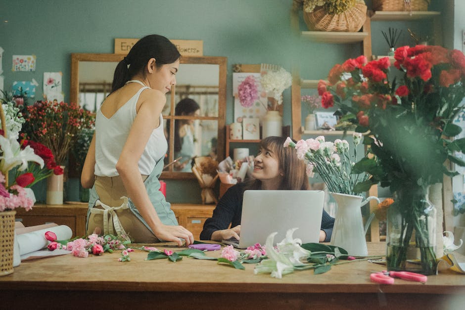 A florist and a customer in a flower shop engaged in a conversation, surrounded by an arrangement of fresh flowers, including pink carnations, white lilies, and red roses. The florist, wearing a white