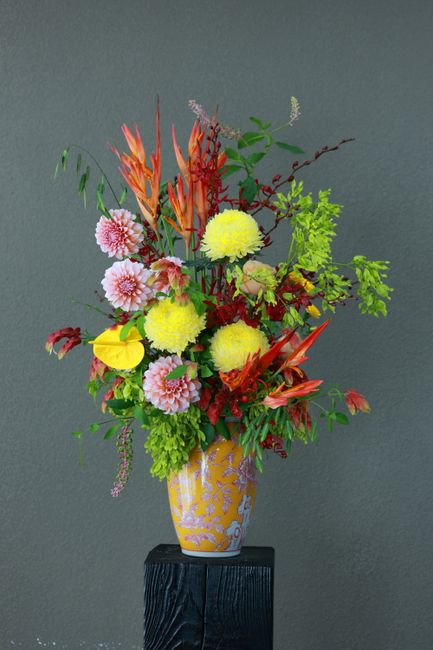 A vibrant floral arrangement in a yellow and white patterned ceramic vase, featuring a mix of flowers including pink chrysanthemums, yellow pom-pom chrysanthemums, and orange-red gloriosa lilies, comp