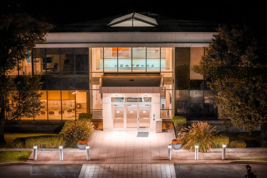 A modern commercial building at night illuminated by interior and exterior lighting, featuring large glass windows revealing an illuminated interior, with a paved entrance path lined with small, illum