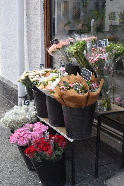 An outdoor display of floral bouquets and arrangements in black buckets, featuring a variety of fresh flowers including pink roses, white and cream blooms, and pink carnations. Some bouquets are wrapp
