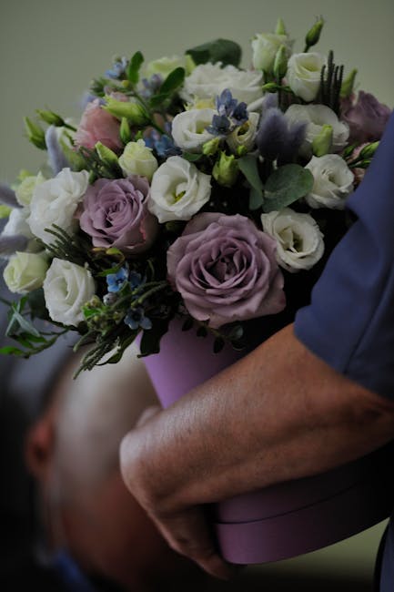 A person wearing a blue sleeve holds a round, pink gift box filled with a floral arrangement featuring white and pale purple roses, small blue flowers, and green foliage. The flowers are fresh, with v