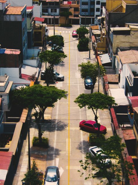 This image offers an aerial view of a residential street lined with multi-story buildings on both sides, with parking spaces along the curb. Several cars, including a prominent pink vehicle, are parke